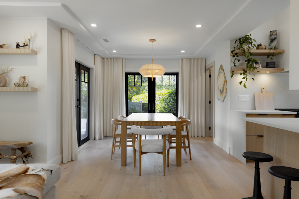 A bright, modern dining space with a wooden table, Scandinavian-style chairs, and large black-framed glass doors leading to an outdoor area.