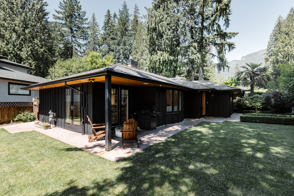 Modern black-clad house with a covered patio, wooden Adirondack chairs, and lush green landscaping, surrounded by tall evergreen trees.