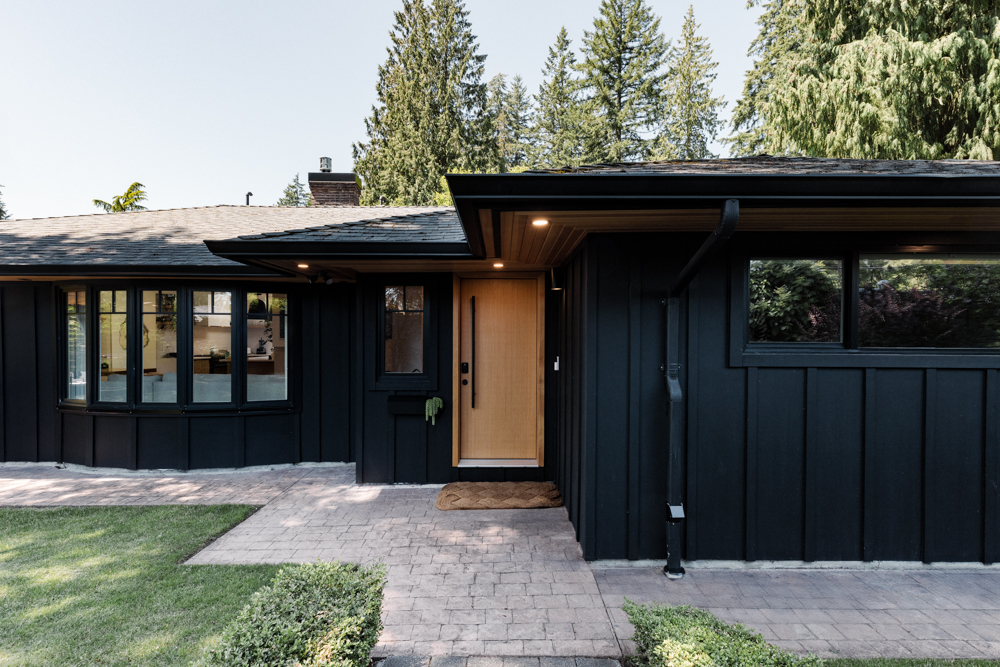 Modern black-clad home entrance with a warm wood front door, brick pathway, and lush greenery.