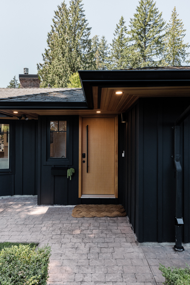 Modern black exterior home with a warm wooden front door, surrounded by lush greenery and illuminated by recessed lighting.