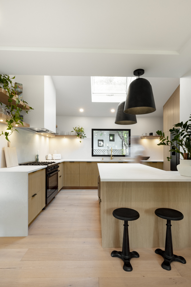 A modern, minimalist kitchen featuring warm wood cabinetry, white countertops, black pendant lights, and a skylight.