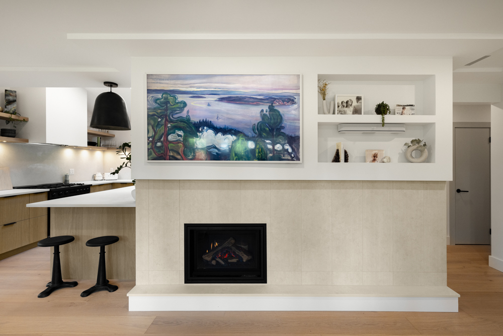 A modern fireplace with built-in shelving and a framed landscape painting, seamlessly dividing the living room and kitchen.